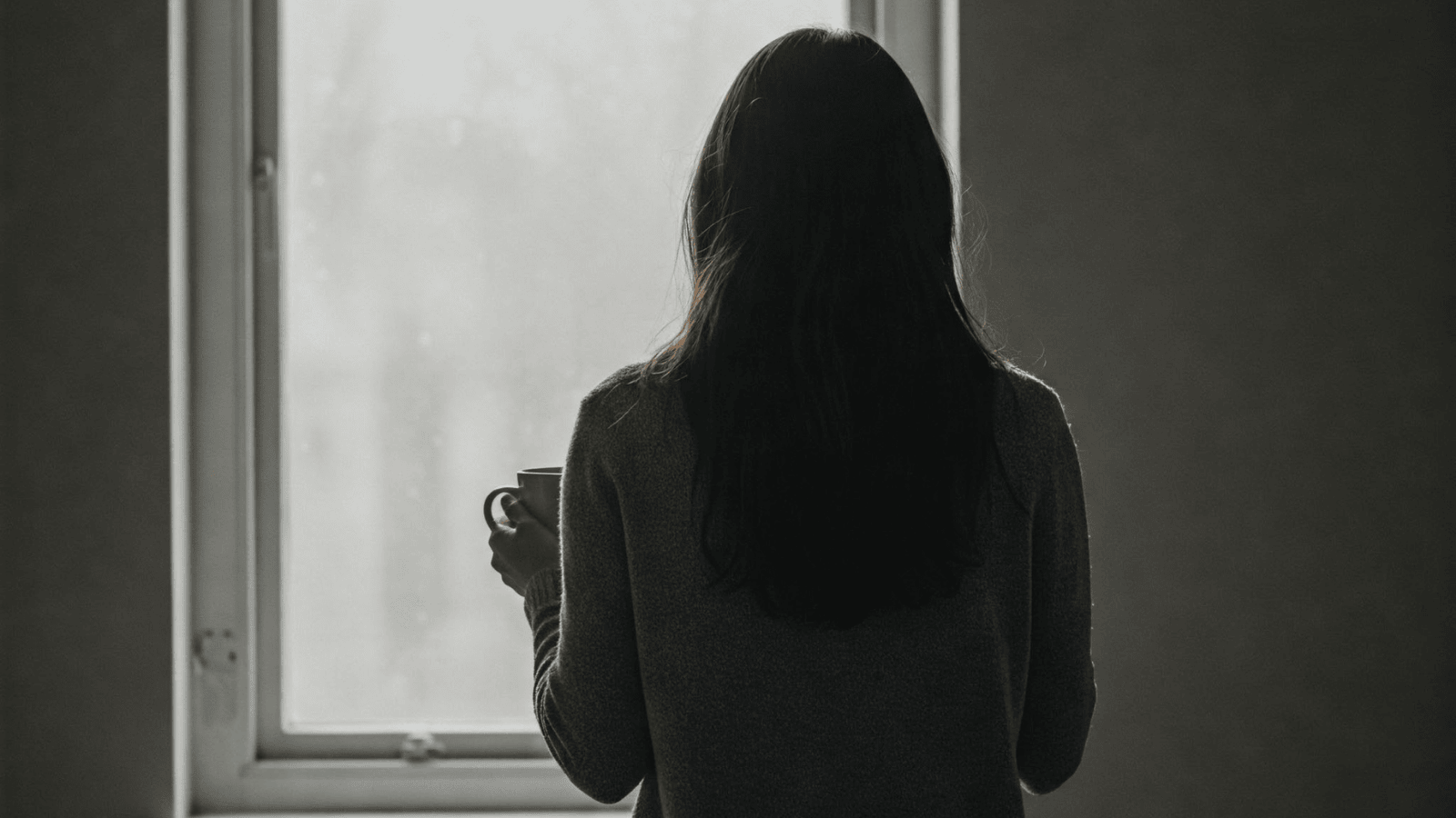 Eine Frau steht mit einer Tasse am Fenster und blickt nach draußen – Symbol für Erschöpfung, Nachdenken und emotionale Überforderung.